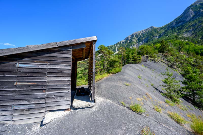 Cabane en bois protgeant le fossile de l'ichtyosaure de La Mlaie  Prads-Haute-Blone, situe sur une pente de marnes grises avec vue sur les montagnes environnantes sous un ciel bleu.